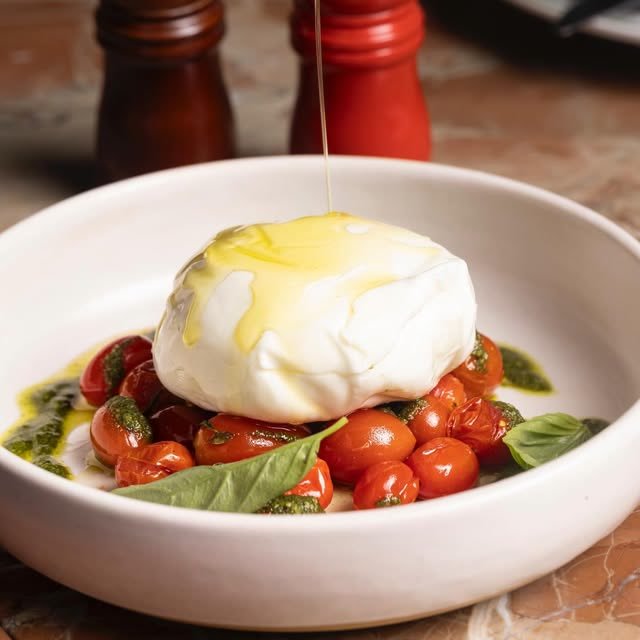 Burrata recebendo um fio de azeite, sobre tomates confit, pesto e folhas de manjericão